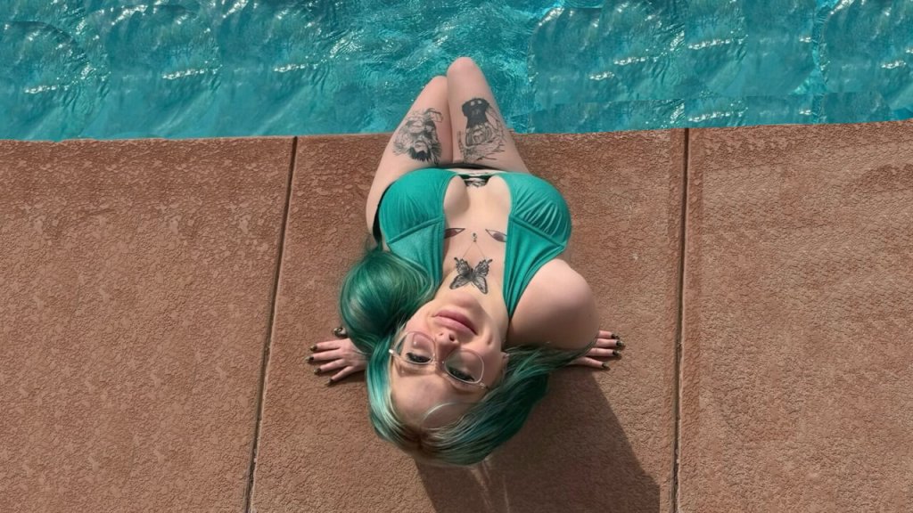 A jassy onlyfans with vibrant turquoise hair relaxing by the pool, wearing a teal bikini and showcasing visible tattoos on arms and chest.