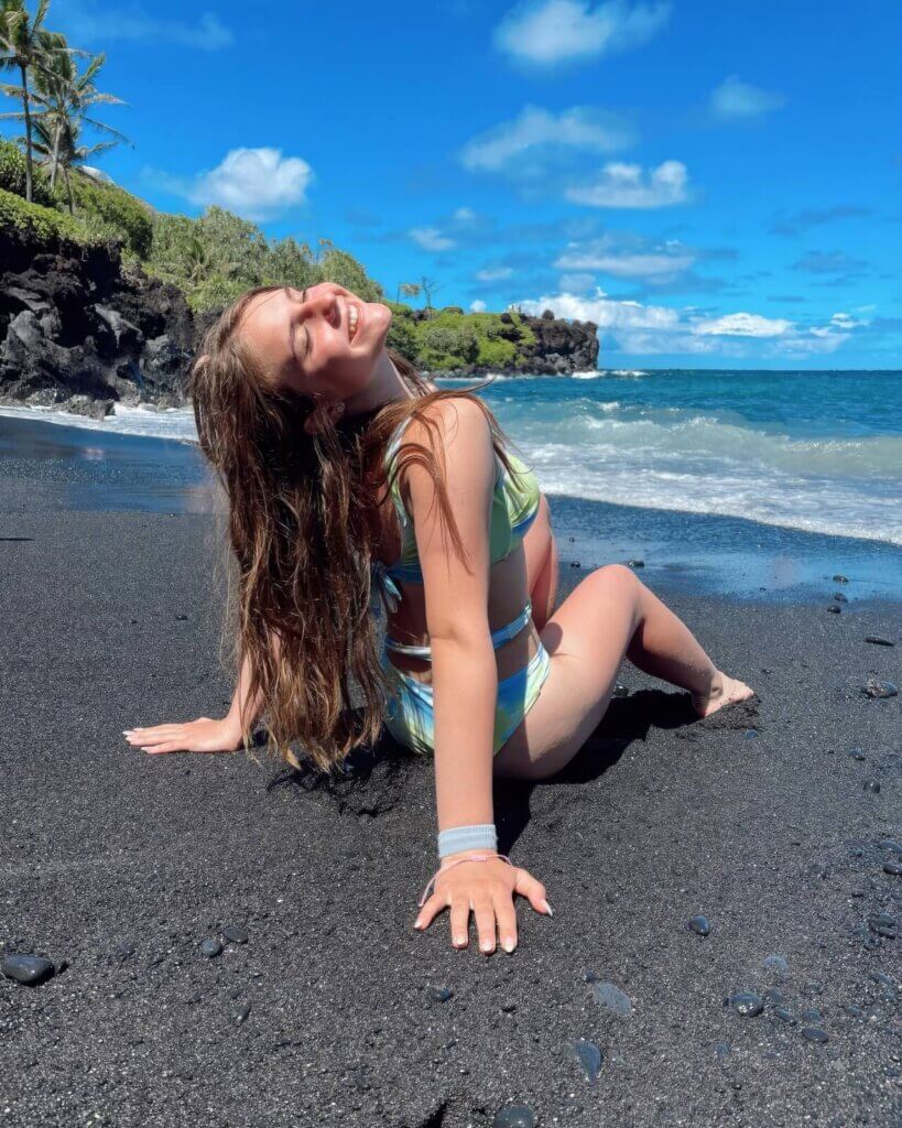 Piper Rockelle sits on a black sand beach, facing the ocean, with green palm trees and a blue sky in the background.