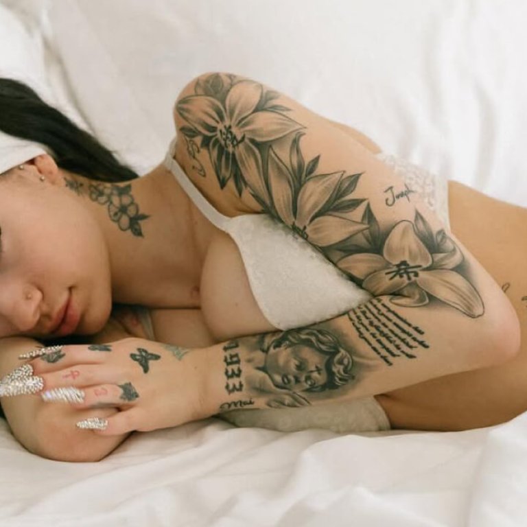 Bhad Bhabie, with intricate tattoos, lies on a white bed in a light bra and pink bottoms, as fans wonder what kind of cancer does bhad bhabie have.