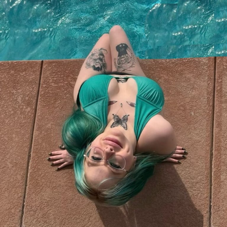 A jassy onlyfans with vibrant turquoise hair relaxing by the pool, wearing a teal bikini and showcasing visible tattoos on arms and chest.