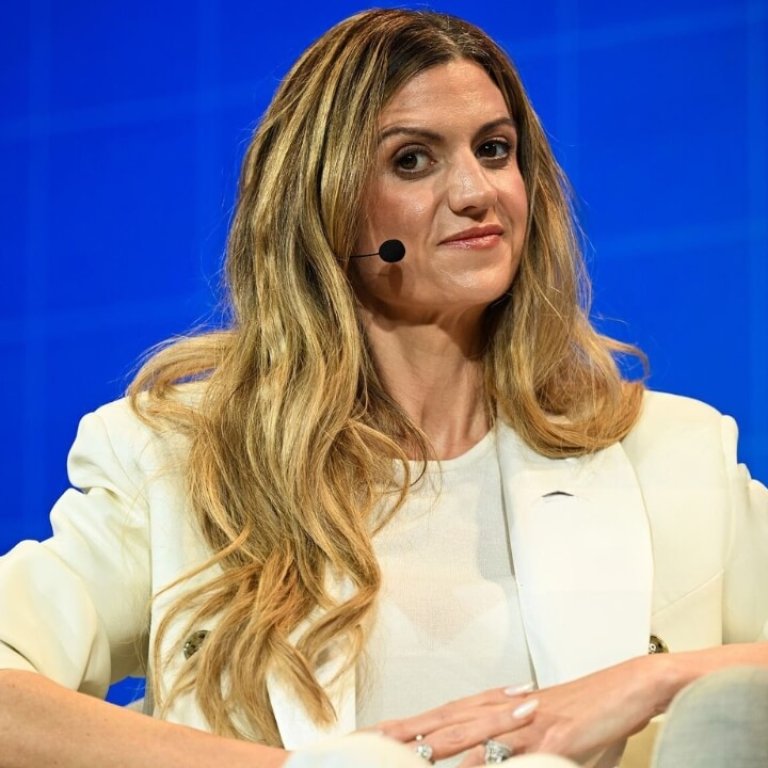 Keily Blair with long, wavy hair sits in a chair on stage, wearing a stylish white outfit against a blue background.